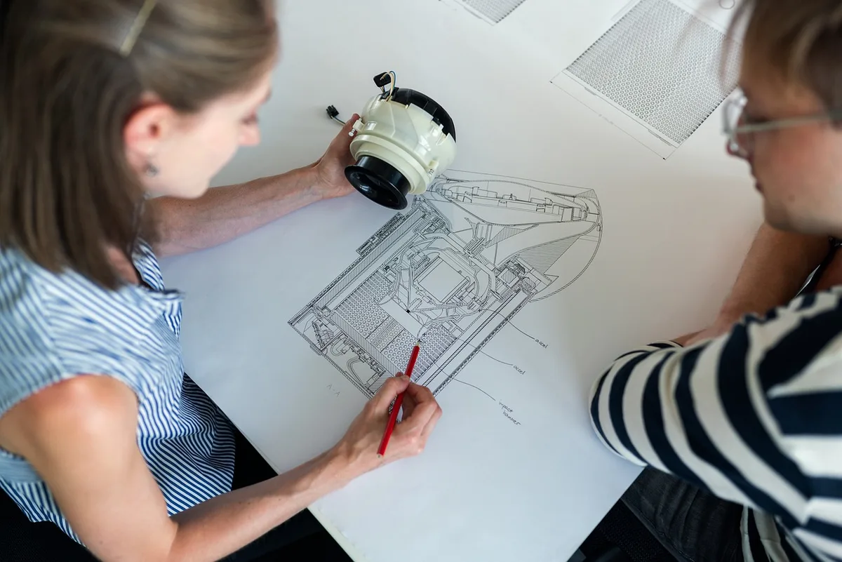 Close-up of hands sketching technical plans beside a laptop and measurement tools, bright interior, anonymous hands