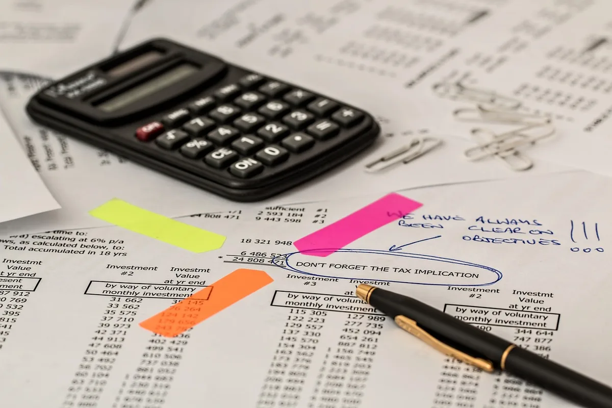 A tidy desk with printed budget pages, a pen, and a closed laptop, hands about to pick up a sheet