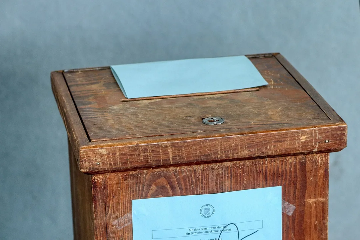 A well-lit desk with hands filling an official election calendar and signing a notice of elections, election forms and a closed envelope visible; anonymous hands only