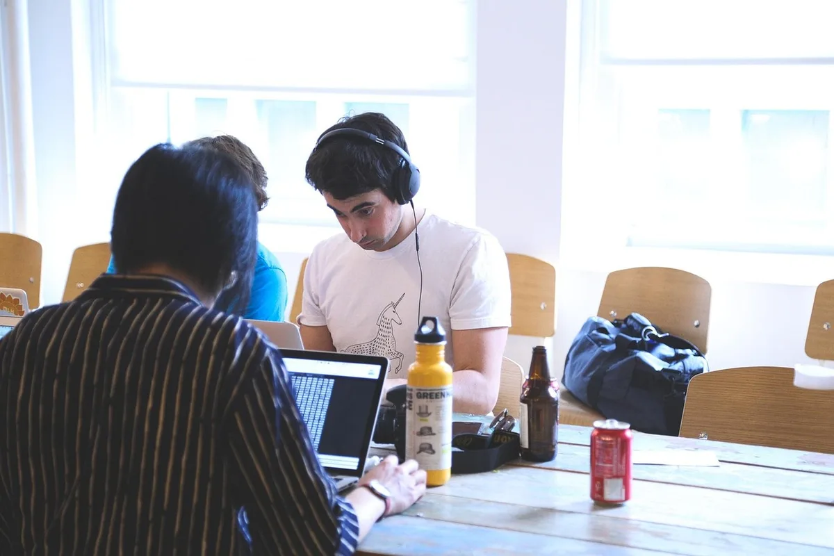 Two anonymous students in a library at a table with laptops and notes, discussing over coffee