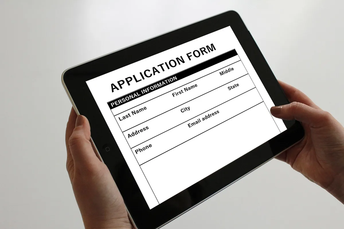 Hands filling an application form on a desk with a laptop and a printed calendar nearby, pen poised above a checkbox